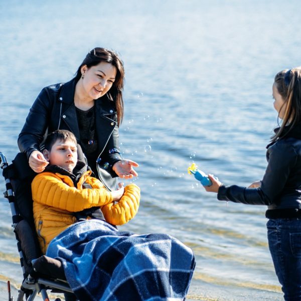 Happy family, mother, daughter and son with cerebral palsy spending time together on the river bank. Teen boy who uses a wheelchair walking with family