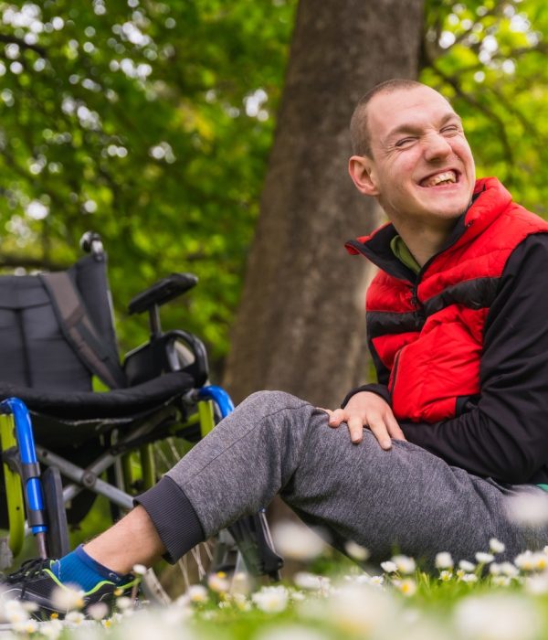 Portrait of a paralyzed young man sitting on the grass next to the wheelchair a daisy flowers, smiling in nature