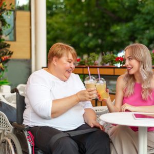 Young daughter and her mother in wheelchair sitting at the table in cafe with drinks and having fun, close up