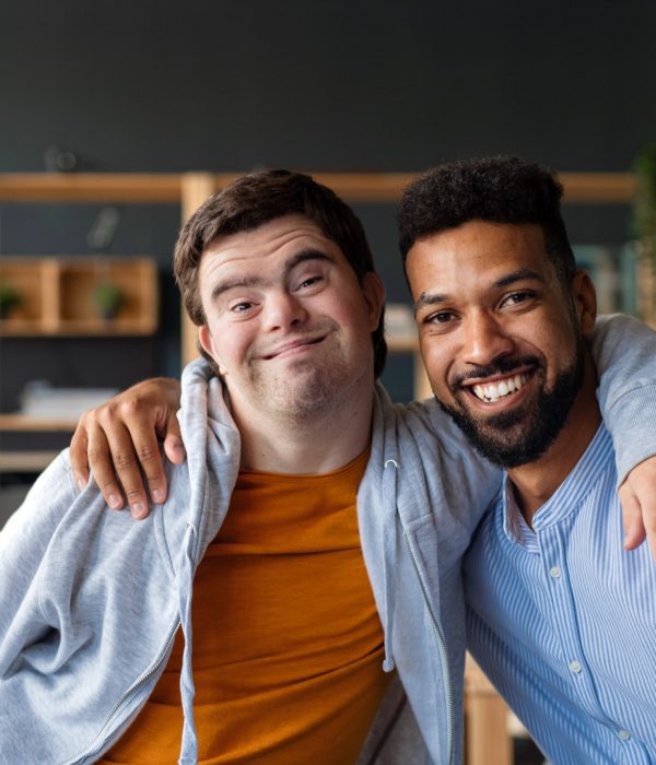 Happy young man with Down syndrome and his tutor with arms around looking at camera indoors at school